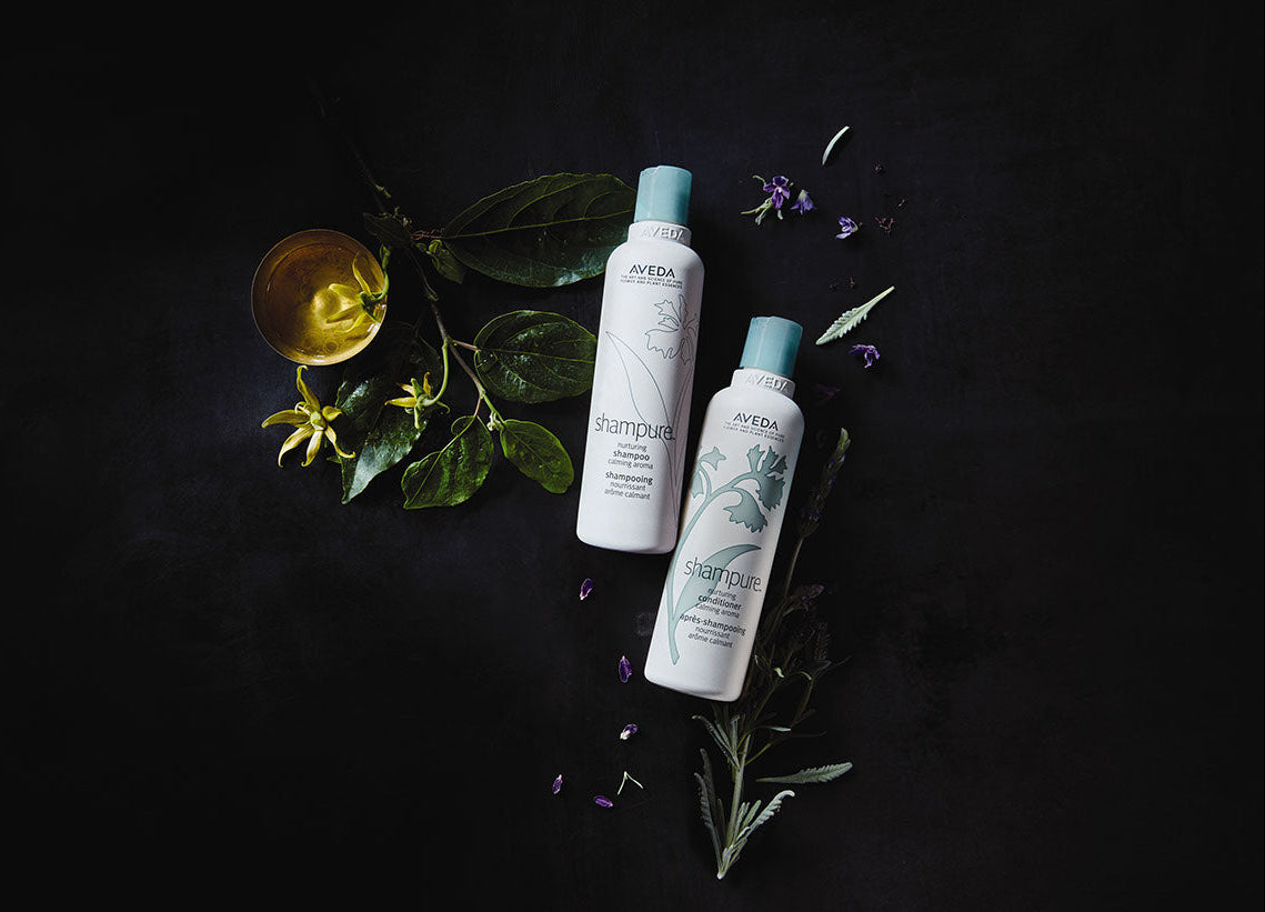 Two bottles of Aveda products on a dark background with leaves and flowers.